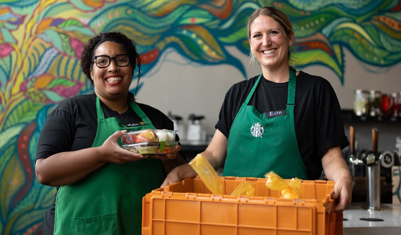 Two women wearing Starbucks green aprons. One holds a protein box and the other an orange FoodShare crate.