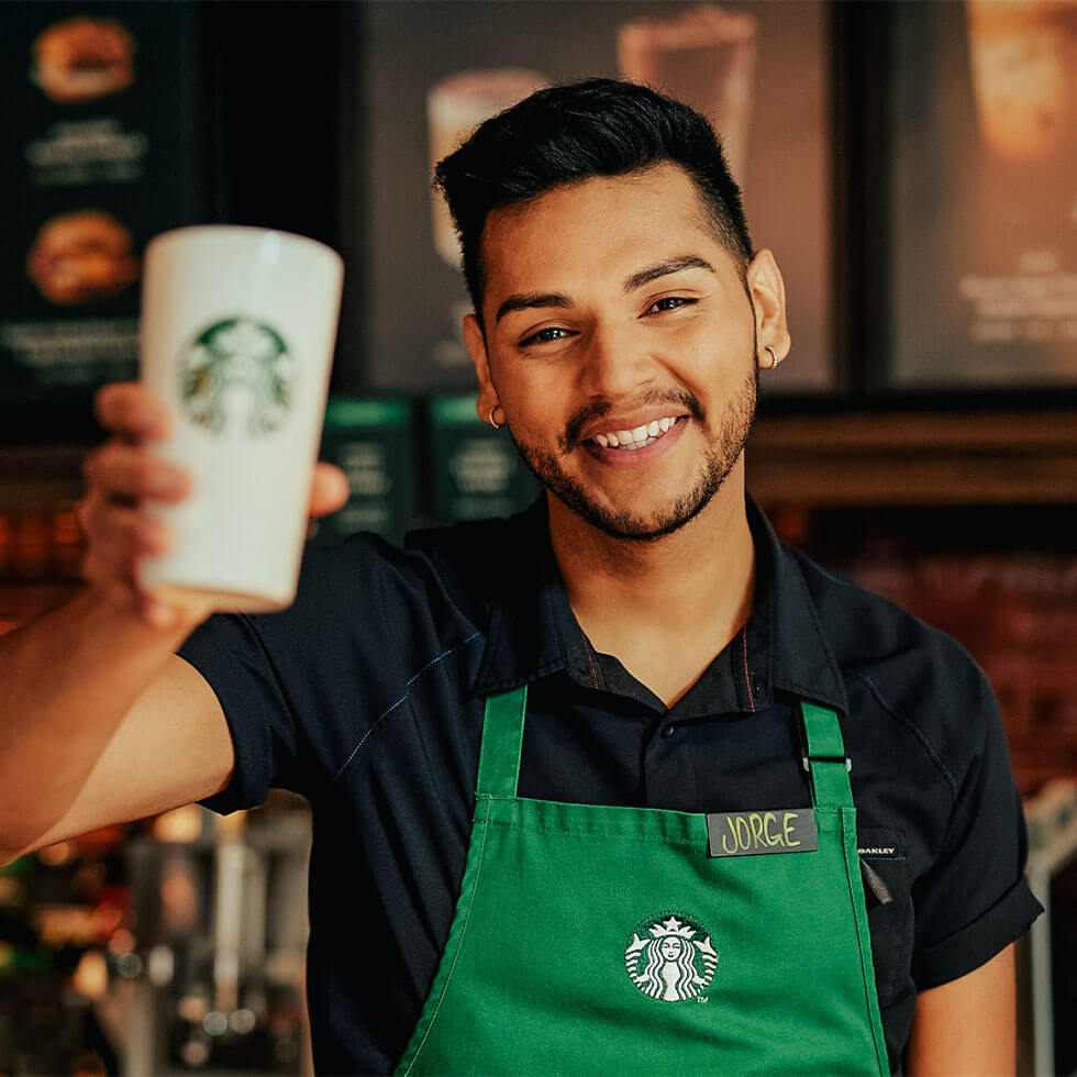 Starbucks barista in a green apron holds up a to-go cup while smiling at the camera.