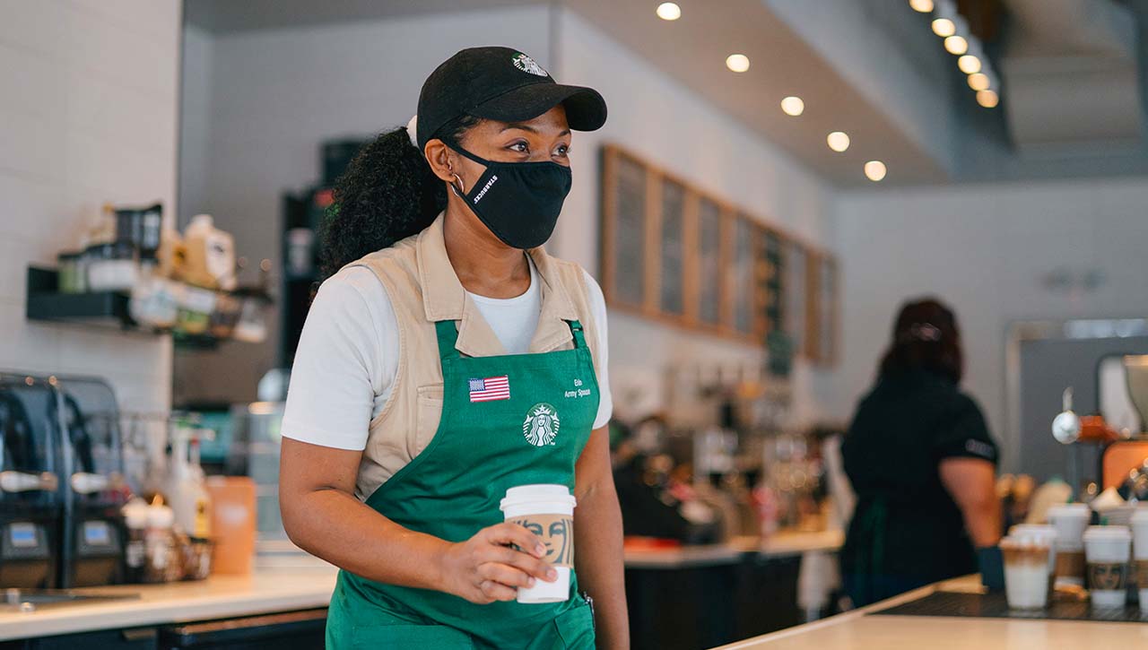 A Starbucks barista behind the counter wearing a green apron and a black facial covering and holding a beverage cup for a customer.