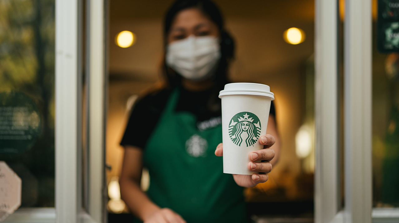 Starbucks partner (employee) in a drive thru window, wearing a green apron and a facial covering, looking at the camera holding a to go cup forwards.