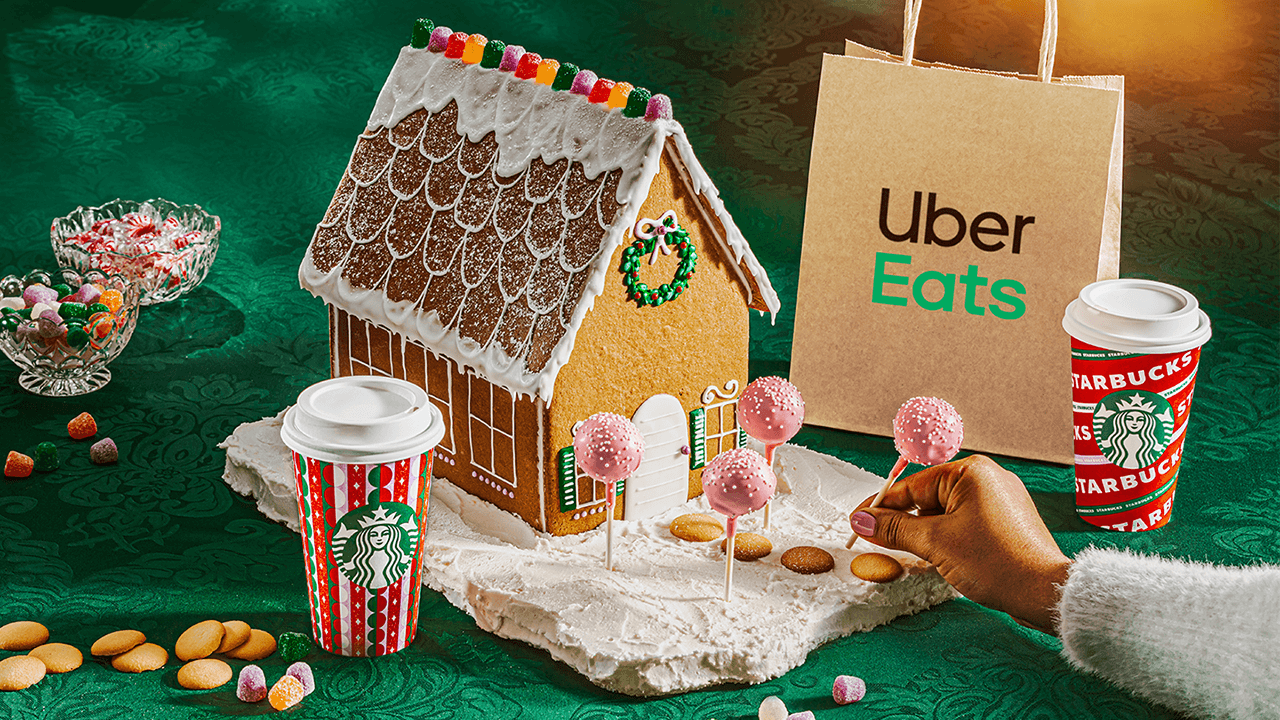 Holiday gingerbread house with Cake Pops whimsically serving as trees in house’s front yard. A hand places the Cake Pops while a Starbucks cup and Uber Eats bag stand on either side.