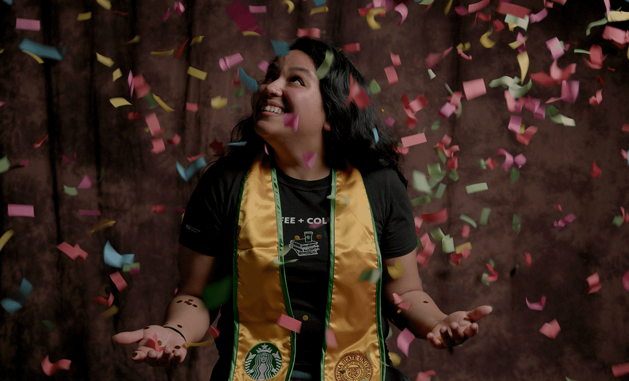 A recently graduated Starbucks partner celebrates under a flurry of confetti. Her gold graduation stole has the Starbucks Siren embroidered on one end and the ASU logo on the other.