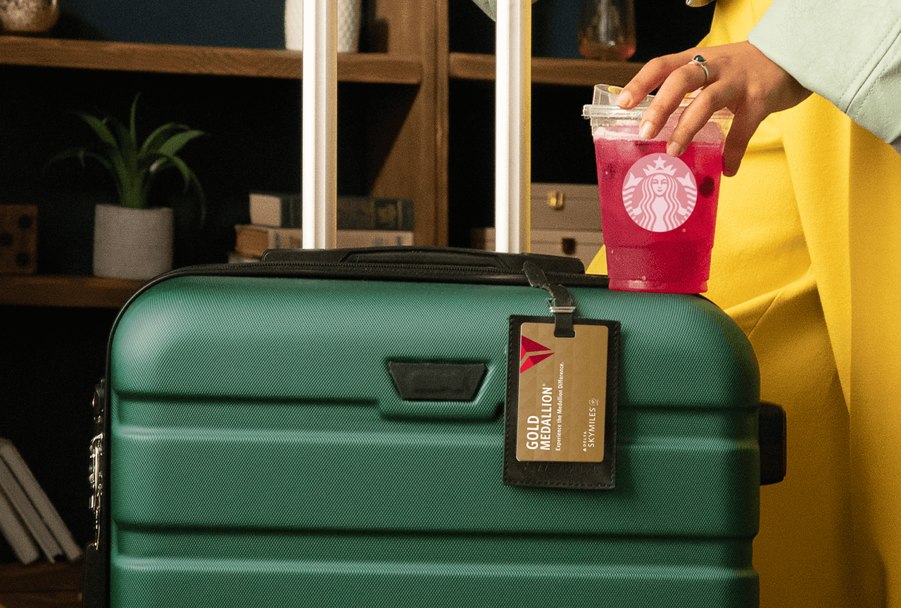 Hand balancing a Starbucks Refreshers&reg; Beverage on a green suitcase with a Delta luggage tag.