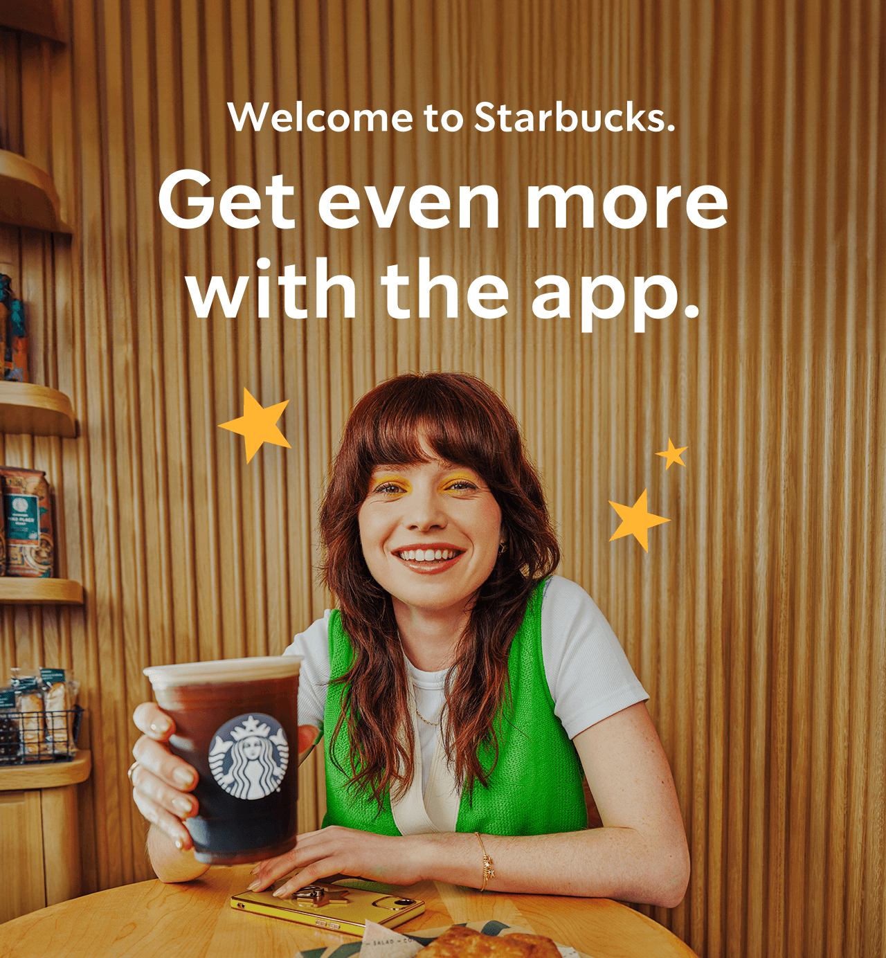 Welcome to Starbucks. Get even more with the app. | A woman in a green vest sitting at a table and holding up a cup of coffee.