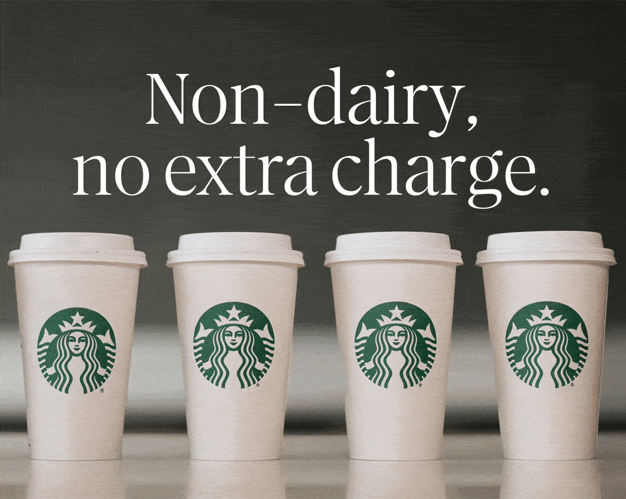 Non-dairy, no extra charge. | Five Starbucks to-go cups sitting on a counter, each turning to reveal a different type of beverage handwritten on the cup; milk, soy beverage, coconut beverage, almond beverage, and oat beverage.