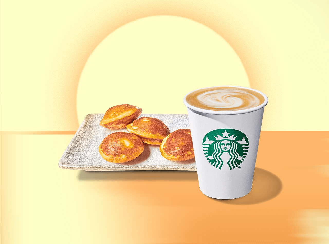 A stoneware plate with four golden brown mini pancakes sitting beside a white Starbucks cup filled with an espresso beverage against a warm yellow background featuring a rising sun.