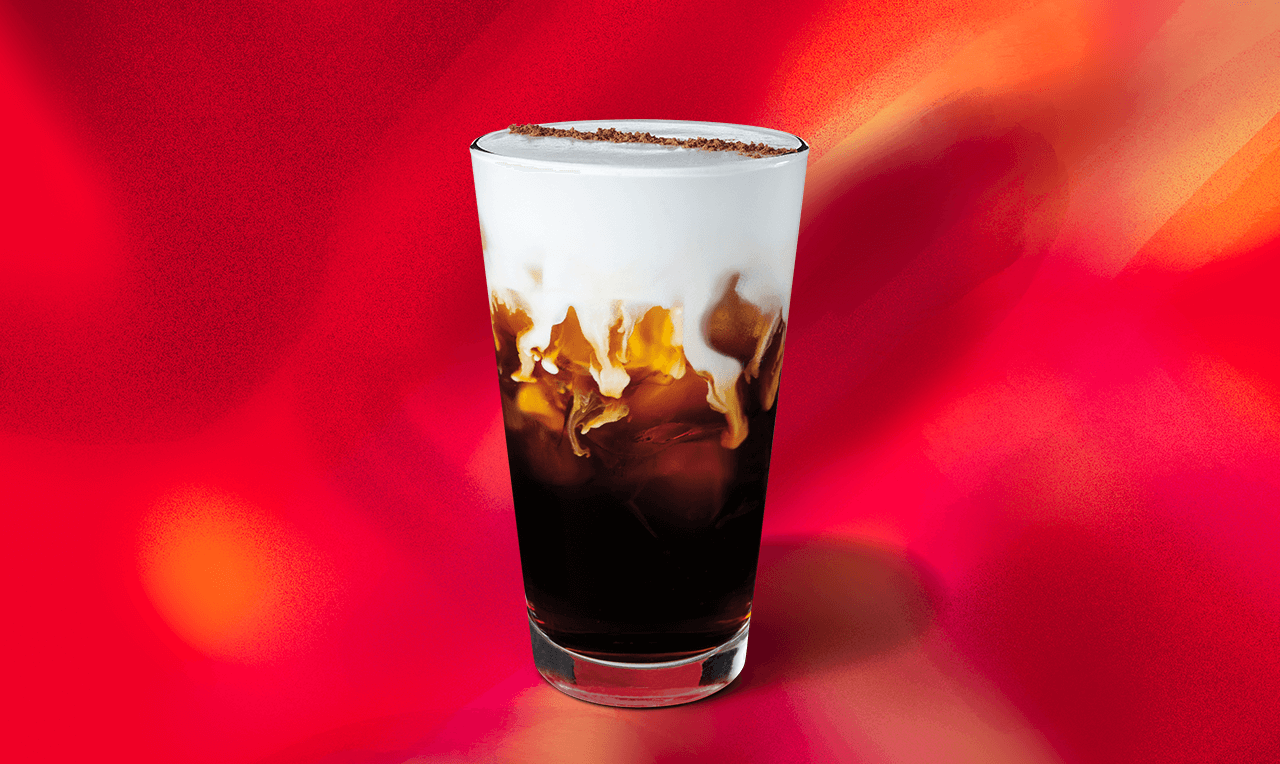 Cold coffee topped with cascading cream in a glass against a red background.