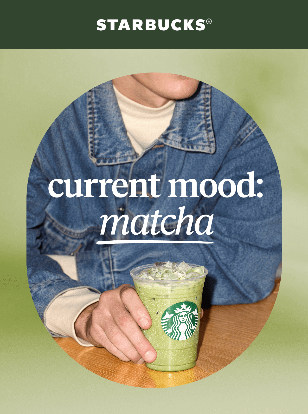 STARBUCKS® | Current mood: matcha | A person in a denim jacket leans over a wooden tabletop while holding a Starbucks iced matcha latte in a to-go cup.