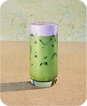 An Iced Lavender Cream Oatmilk Matcha sitting on a tabletop with a sage green backdrop.