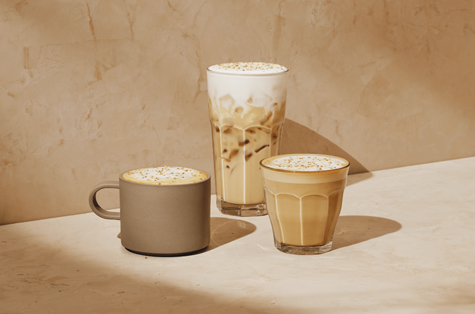 3 espresso drinks are sitting against a light beige background. On the left, a latte topped with rich foam and sprinkled with crunchy sugar. Next to it, we see a tall creamy iced latte, beige in colour with thick cold foam on top. On the far right, we see a cortado beautifully layered with rich espresso and cream.