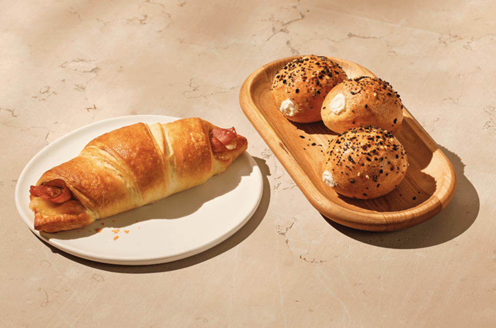 2 savoury bakery snacks lie on a warm beige background. On the left, a freshly warmed flaky ham & swiss croissant. Next to it is a trio of golden mini bagel rounds topped with an assortment of seeds and seasoning.