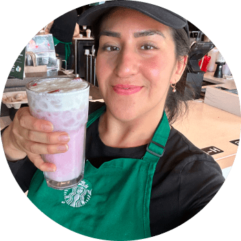 Circular image of barista Antonella wearing a green apron and black hat, holding a Pink Drink with Vanilla Sweet Cream Cold Foam in a glass.