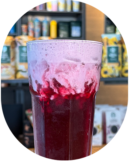 A Blackberry Sage Refresher with Lavender Cream Cold Foam in a glass placed on a Starbucks countertop in front of packaged coffee on shelves.