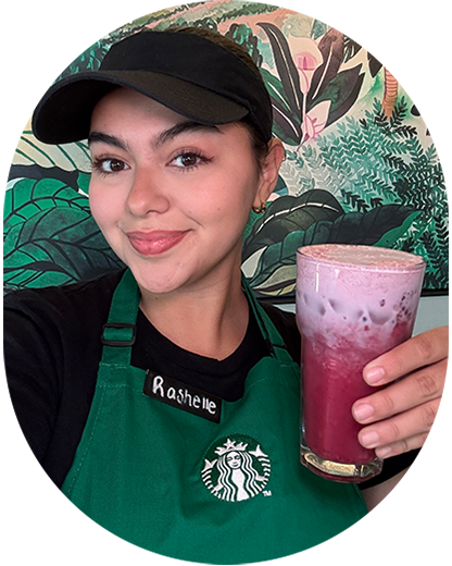 Barista Rashelle, wearing a green apron and black visor, holds a Midnight Drink with Lavender Cream Cold Foam in a glass. She stands in front of a wall painted with lush foliage.