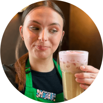 Circular image of barista Maddie wearing a green apron and name tag, holding an Iced Vanilla Latte with Cherry Cream Cold Foam in a glass.