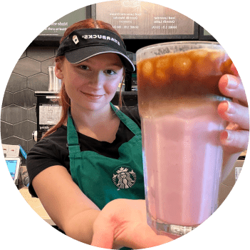 Circular image of barista Maeve wearing a green apron and black Starbucks visor, holding an Iced Cherry Macchiato in a glass.