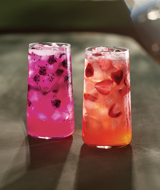 There’s a photograph of a Starbucks Mango Dragonfruit and Strawberry Açaí Lemonade Refresher®. One is orange and the other is pink—both have ice and fruit inclusions. Sunlight is shining off both glasses.